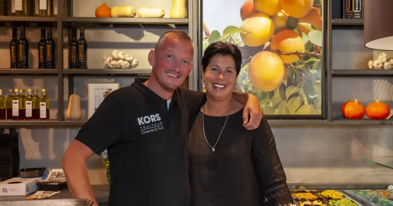 Twee medewerkers van Kors lachend in de winkel, voor een schap met olie, pompoenen en een foto van sinaasappels